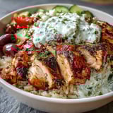 A finished Greek Chicken Bowl topped with creamy tzatziki, briny Kalamata olives, fresh herbs, and lemon wedges.