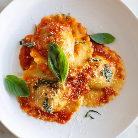 Golden spinach ricotta ravioli on a dinner plate, drenched in rich red marinara and topped with fragrant basil leaves.