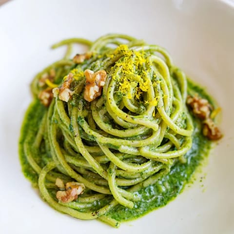 A close-up of creamy spinach walnut pasta in a white bowl, topped with fresh lemon zest and cracked pepper.